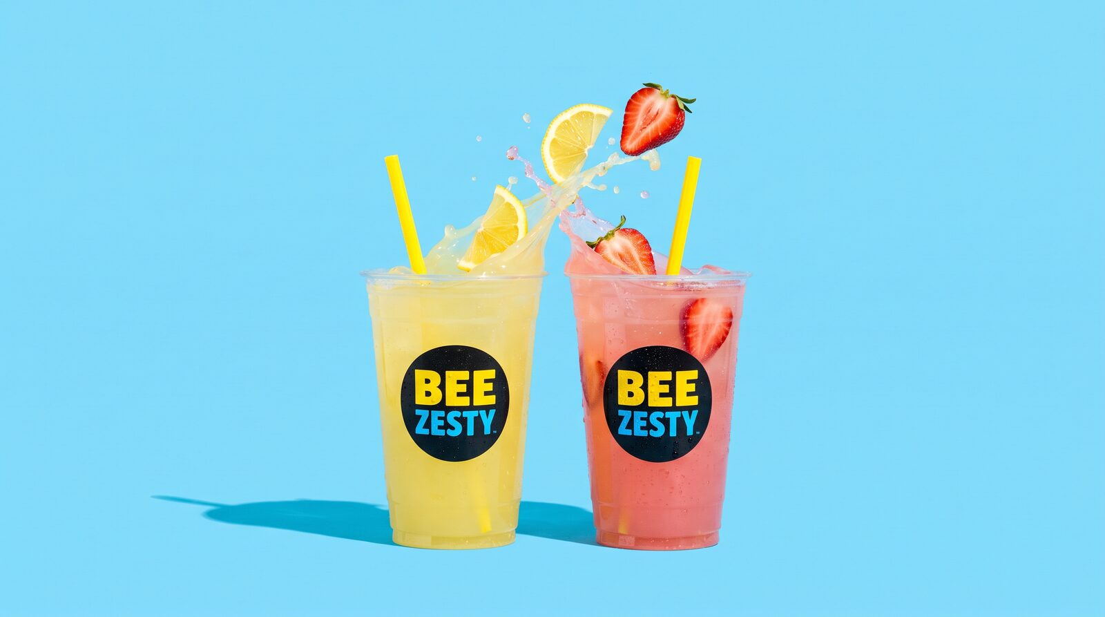 Two icy 32oz BeeZesty cups — classic lemonade and strawberry lemonade — with a splash of juice and flying fruit